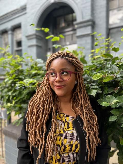 Headshot of Namina Forna wearing red glasses, a black jacket, and a yellow, black, and white blouse. Her hair is brunette and long, and she is looking up at a corner and smiling. The background shows a grey building and green plants.