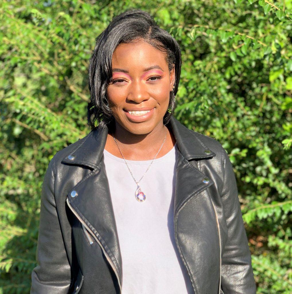 Headshot of Nia Davenport in a black leather jacket and white top. Her black hair is short, and she is wearing pink makeup and a colorful necklace. The background is out of focus but shows plants. She is smiling at the camera.