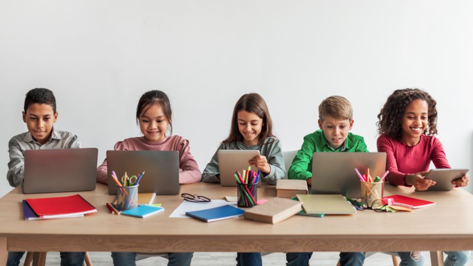Five kids are sitting at a desk, using tablets and computers.