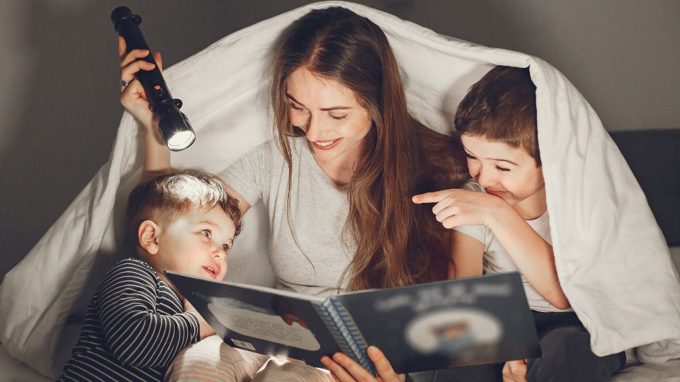 A mom and two sons are reading in the dark with a flashlight. They have a white blanket draped around them making a little tent.