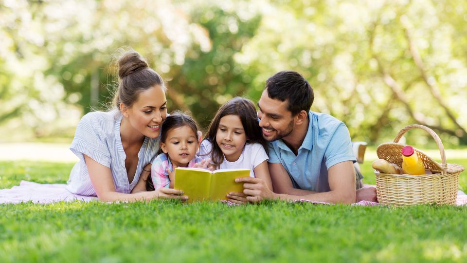Mom and dad reading with their two daughters on a picnic blanket in the grass.