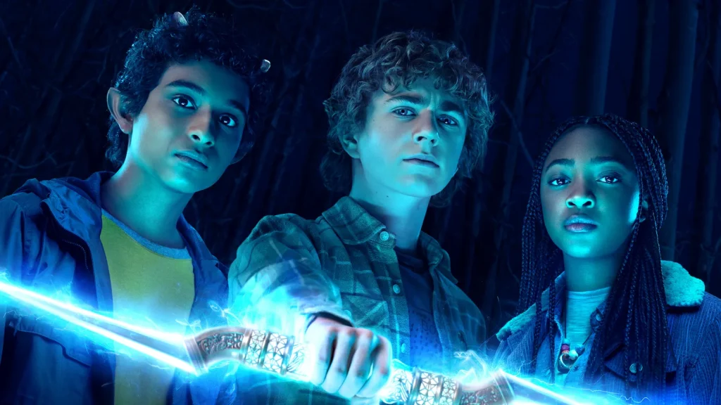 A promotional still for the TV show Percy Jackson and the Olympians. Three pre-teens stand bathed in electric blue light with determined expressions. On the left, is a boy with brown skin, curly black hair, small horns, and pointed ears. In the middle, is a boy with pale skin and curly blonde hair. He grips a lightning bolt from a central, carved hilt. On the right, is a girl with dark brown skin and black hair in multiple small braids.