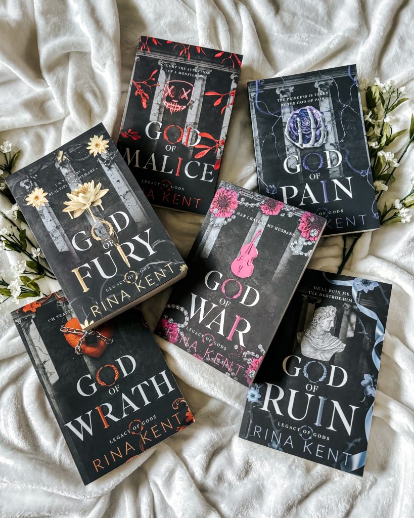 On top of a cozy white blanket and two bundles of white flowers sits the black, Greek mythology inspired covers of Anna Kent's Legacy of Gods series.