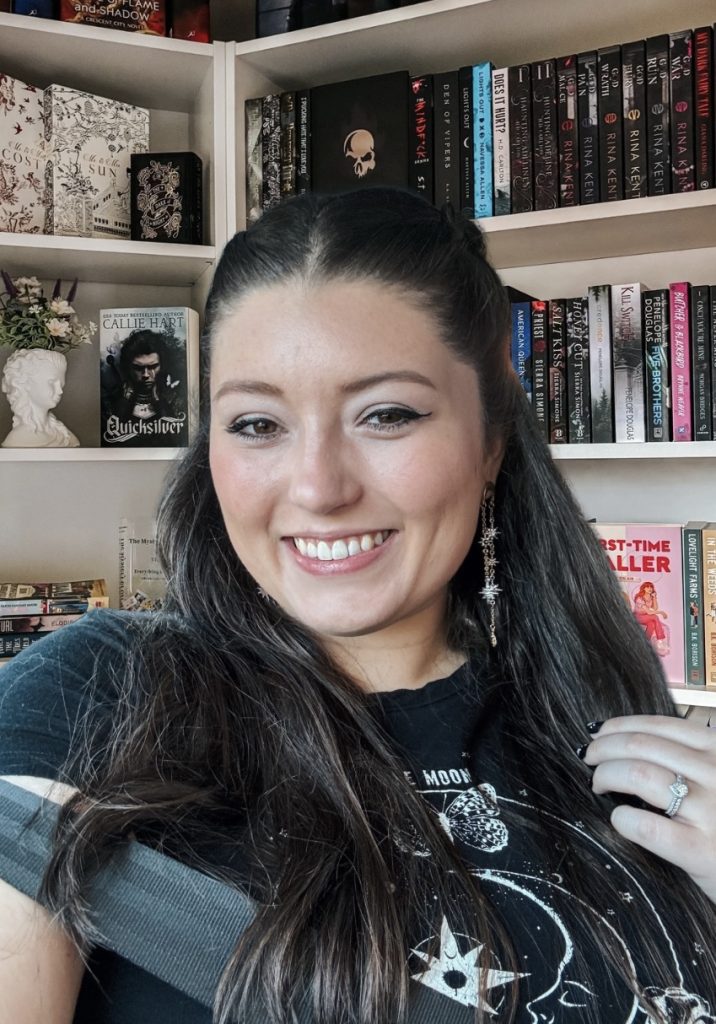 Bookstagrammer Makenna smiles in front of a white bookcase with dark-colored book bindings behind her. Makenna is wearing a black t-shirt, dangly star earrings, and black winged eyeliner.