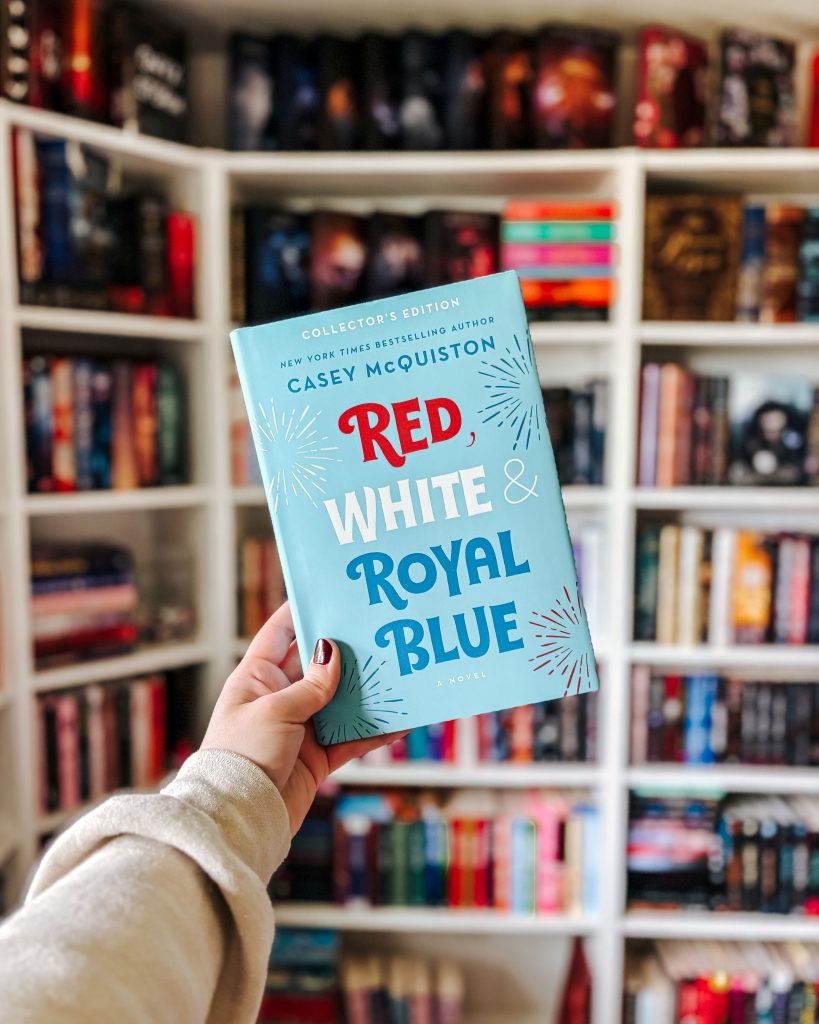 Makenna's hand, complimented with burgundy nail polish, holds up the romance novel Red, White, and Royal Blue by Casey McQuiston. The cover is a light blue with red white and blue fireworks. The book is in front of a blurred out bookcase stacked with novels.