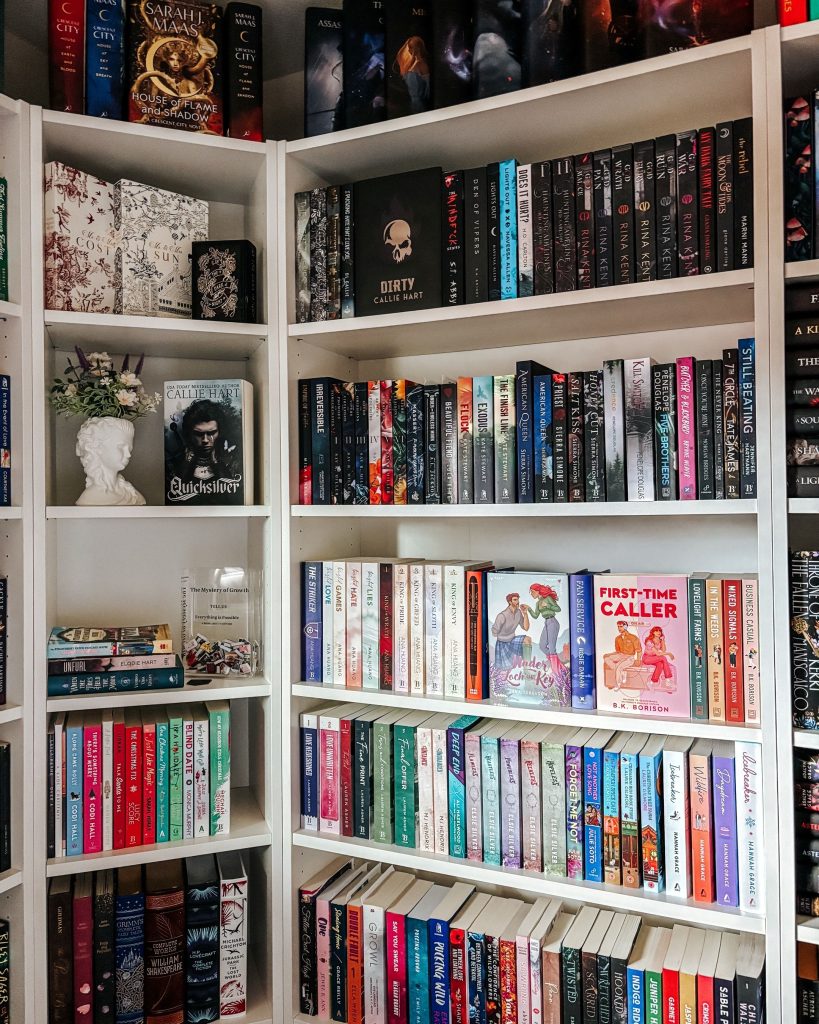 Bookstagrammer Makenna's personal white bookshelf. In the corner sits a bust of a woman's head with a bouquet of flowers coming out of it; on the remaining shelves are various novels. The top two shelves have dark book bindings showing and the bottom two have light and colorful bindings.