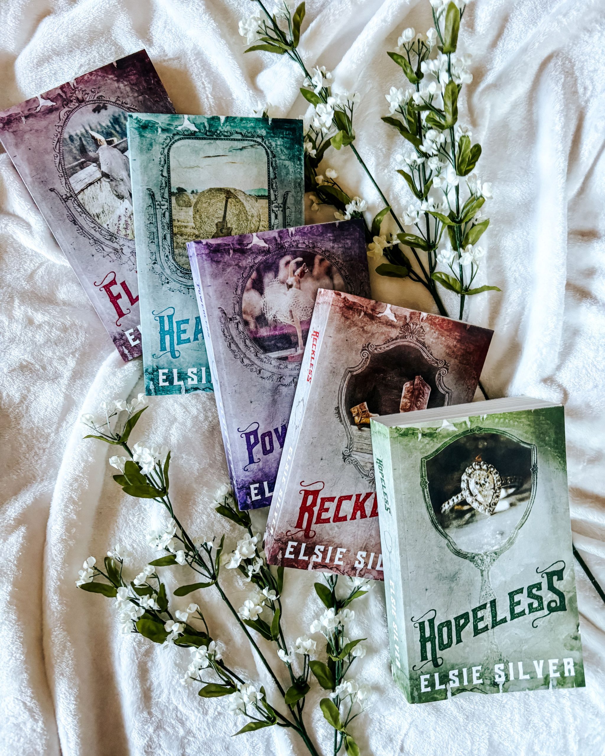Elsie Silver's Chestnut Springs Series. Four of Silver's colorful, but faded, covers splayed over one another with Hopeless by Elsie Silvers on top. To the left and right of the books are leafy, white budded flowers.