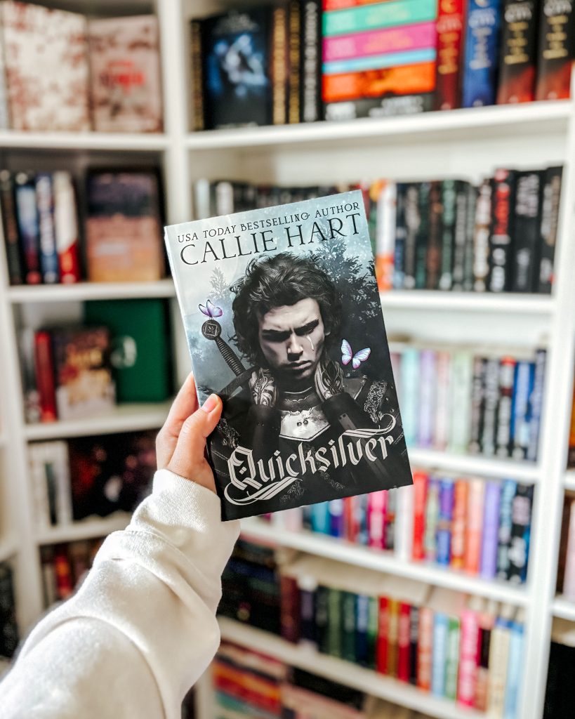 Makenna holds up the romance, fantasy book cover Quicksilver by Callie Hart. The cover features the character Kingfisher, suited in armor, looking down with a single tear falling on his face. In the background is Makenna's blurred out book case.