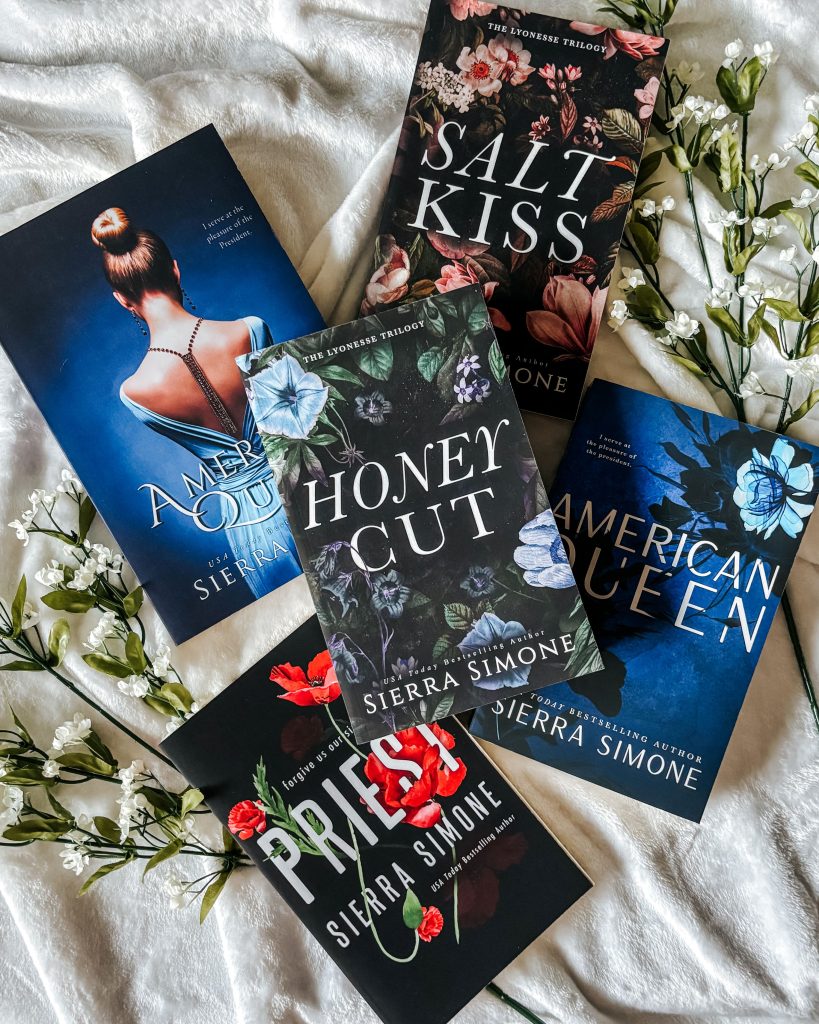 Five Sierra Simone novels on white sheets with white, leafy flowers off to the side. Four novels (Salt Kiss, two copies of American Queen, and Priest) are in a circle-like orientation with the novel Honey Cut resting in the center of them.