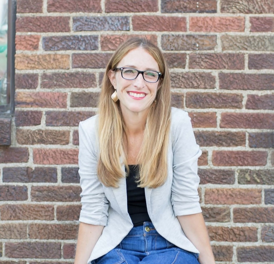 Headshot of Jaime Sumner, a pale blonde woman with black glasses. She is wearing a grey cardigan, black top, blue jeans, and gold earrings. She is sitting down smiling, and the background behind her is a brick wall.