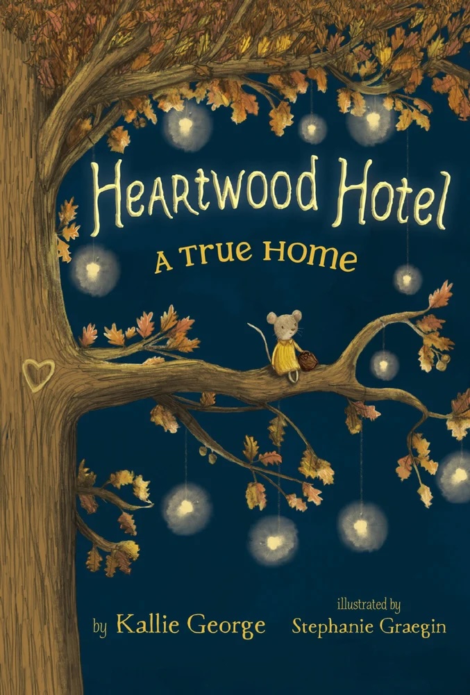 Heartwood Hotel: A True Home by Kallie George, illustrated by Stephanie Graegin, depicting a small grey mouse in a yellow dress holding an acorn while sitting on a tree branch. The trunk of the tree is on the left side of the book, and the branches are full of orange leaves and hanging yellow lights. The background is navy blue.