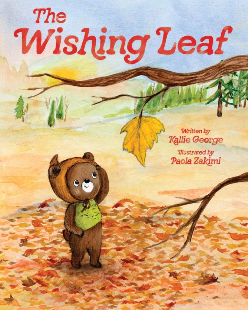The Wishing Leaf written by Kallie George and Illustrated by Paola Zakimi, depicting an illustration of a small brown bear in a brown hat and green shirt looking up at the only remaining yellow leaf on a tree branch. The background features fall leaves, trees, and the setting sun. The title is in red letters at the top of the book cover.