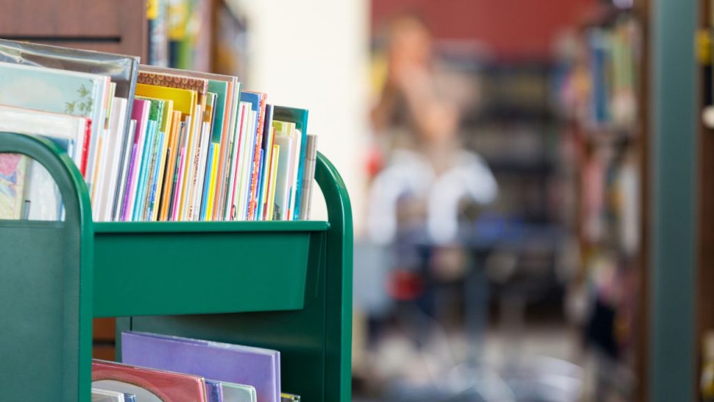 Green cart filled with colorful books in a library setting. Blurred background with bookshelves and indistinct person.