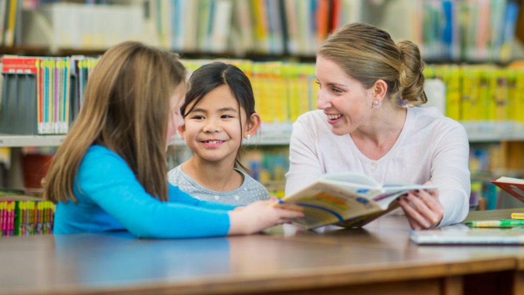 A woman reads a book with two young girls in a library. They are seated at a table, surrounded by bookshelves, and smiling.