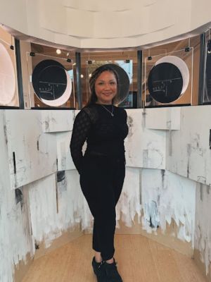 Black paranormal romance author, Britney S. Lewis smiling sweetly at the camera. She is wearing black clothing, has her hair tied back, and is posing in front of a display for an author event.