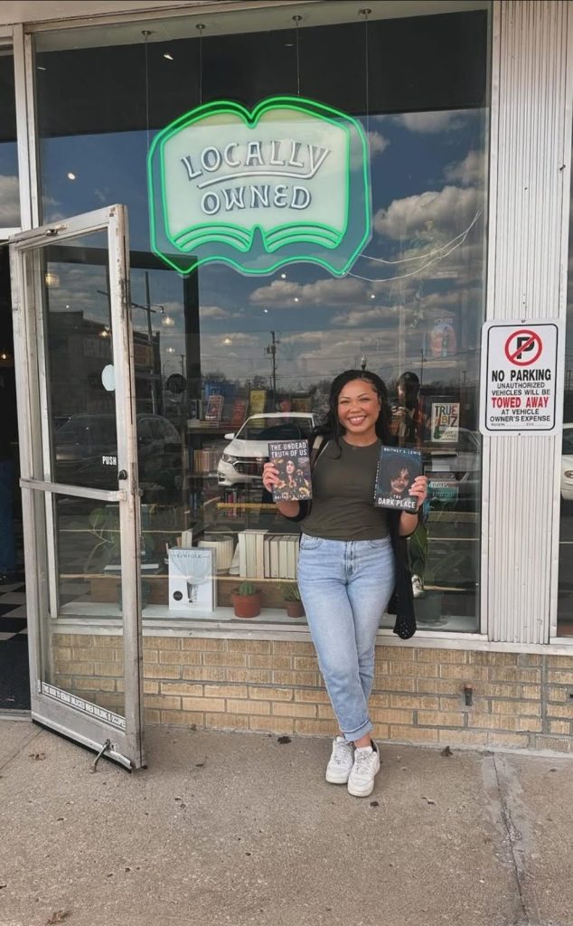 Britney S. Lewis, paranormal romance author, a Black woman wearing jeans and a green top, standing outside an independent book shop, holding two of her published books.