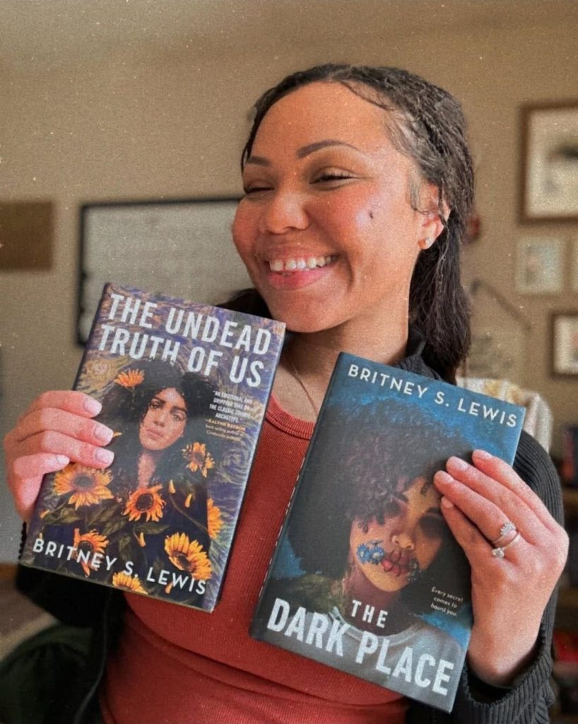 Britney S. Lewis, paranormal romance author, a Black woman with hair up, smiling whilst holding her two published books 'The Undead Truth of Us' and 'The Dark Place'
