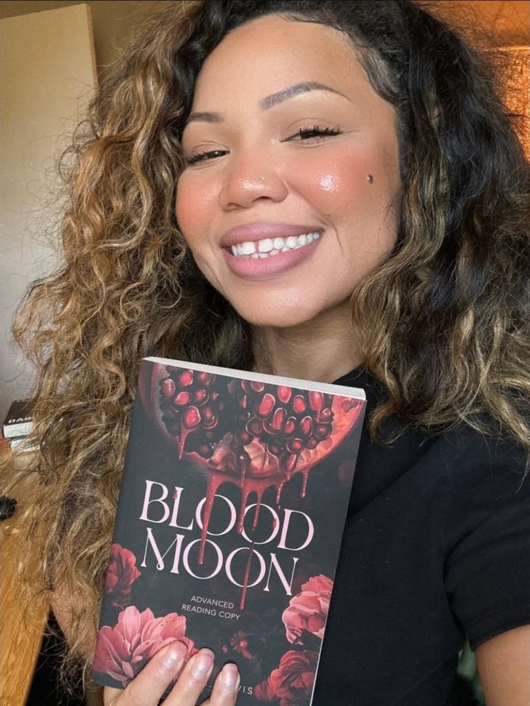 Britney S. Lewis, a paranormal romance author. A Black woman with curly hair smiling fully. Wearing a black top and holding up an Advanced Reading Copy of her book Blood Moon.