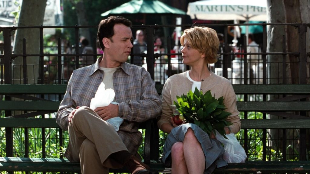 Still from You've Got Mail depicting Kathleen Kelly (Meg Ryan) with short hair in a beige cardigan and blue skirt holding a plant and Joe Fox (Tom Hanks) in a flannel and beige slacks holding a bag. They are sitting on a park bench looking at each other.