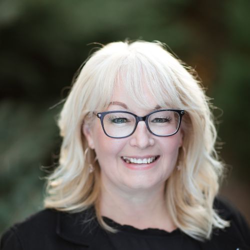 Rom-com author Lynn Painter, a light skinned blond haired woman in black framed glasses and blouse smiles at the camera while outdoors.