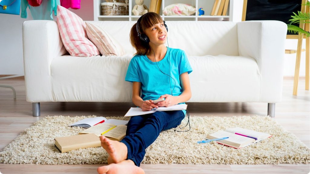 A tween girl sitting on the floor of a living room in front of a couch, on a rug with headphones on while books are spread out around her on the floor.