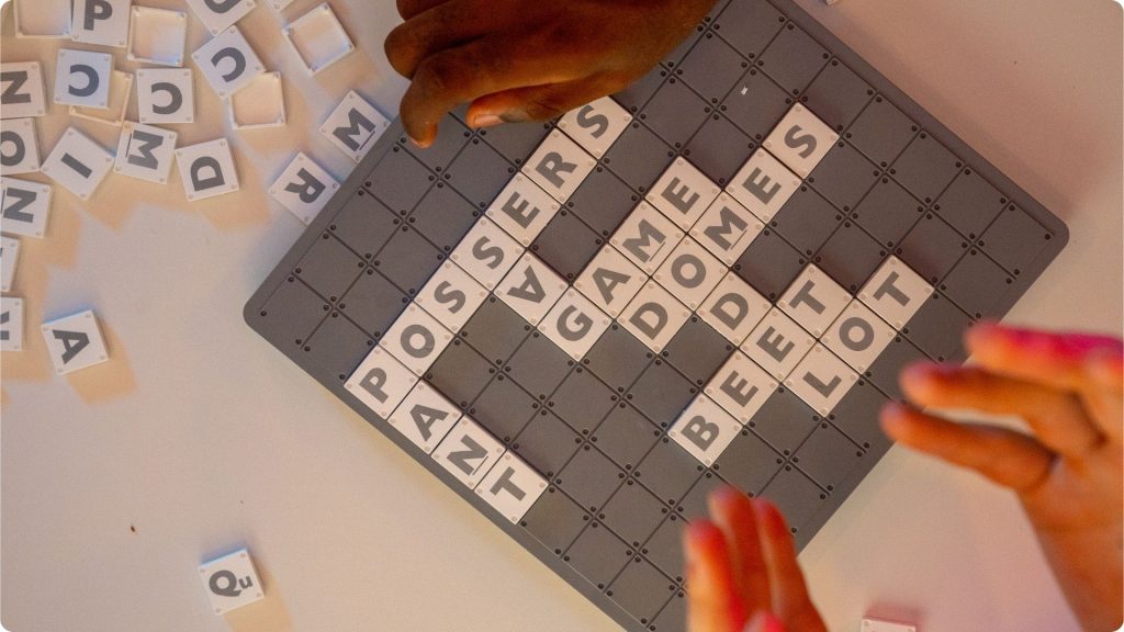 A Scrabble game board sitting on a table with some letter spilled off to the side and hands shown above and to the side.