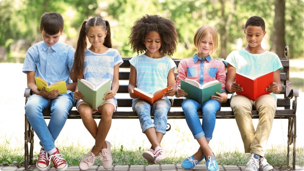 Five children from different cultures are sitting on a park bench outside, each with a different book in their lap.