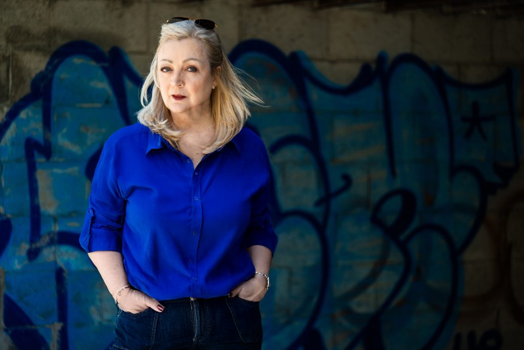 Author Ramona Ballie, a light-skinned blond woman wearing a navy blouse and jeans stand with her hands in her pockets before a graffitied wall.
