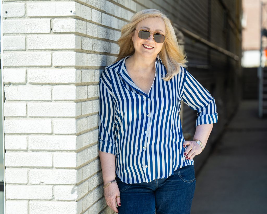 Author Ramona Baillie, a light skinned blond woman wearing a blue and white vertical stripped shirt leans against a pale brick building.