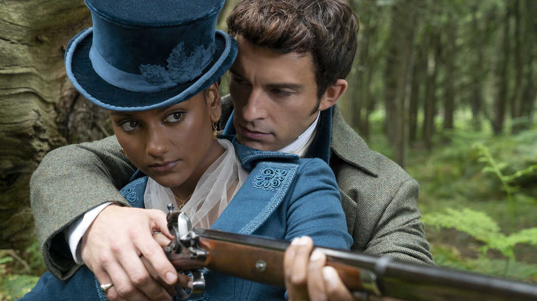 Still from Bridgerton depicting Jonathan Bailey as Anthony Bridgerton and Simone Ashley as Kathani Bridgerton. She is holding a hunting gun, and he is wrapped around her to help her aim. He is looking at her while she is looking off into the distance.