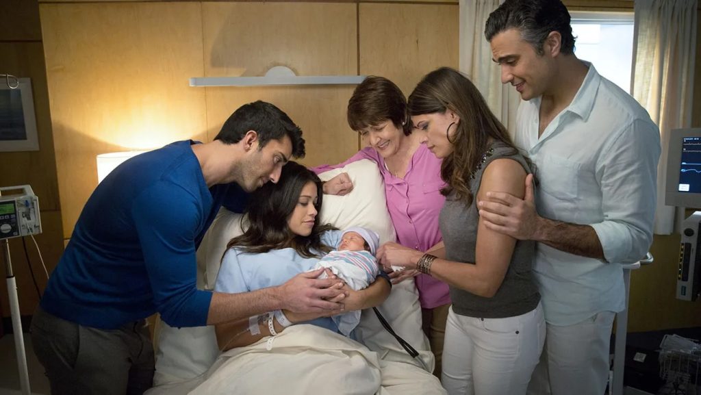 Still from Jane the Virgin depicting Jane Villanueva (Gina Rodriguez) in a hospital bed looking down at the newborn baby in her arms. Surrounding the hospital bed are two women and two men looking down at the baby.
