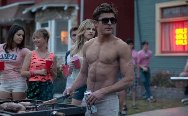 Still from Neighbors depicting Zac Efron as Teddy at an outdoor frat party. He is shirtless and cooking something on a grill. Behind him, there are people with red solo cups looking in the same direction as him.
