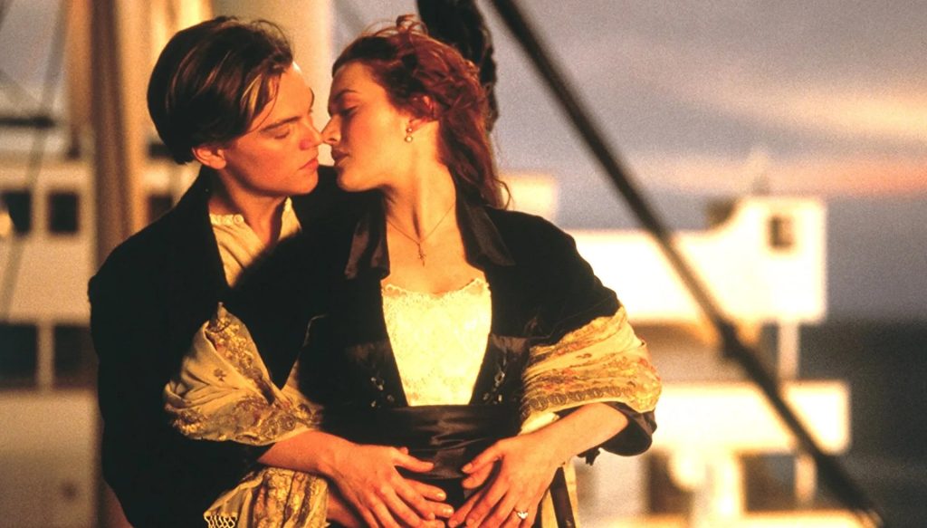 Still from Titanic featuring Leo DiCaprio and Kate Winslet with their faces close together as if they are about to kiss. The background is blurry but depicts parts of a boat, the sky, and the ocean.