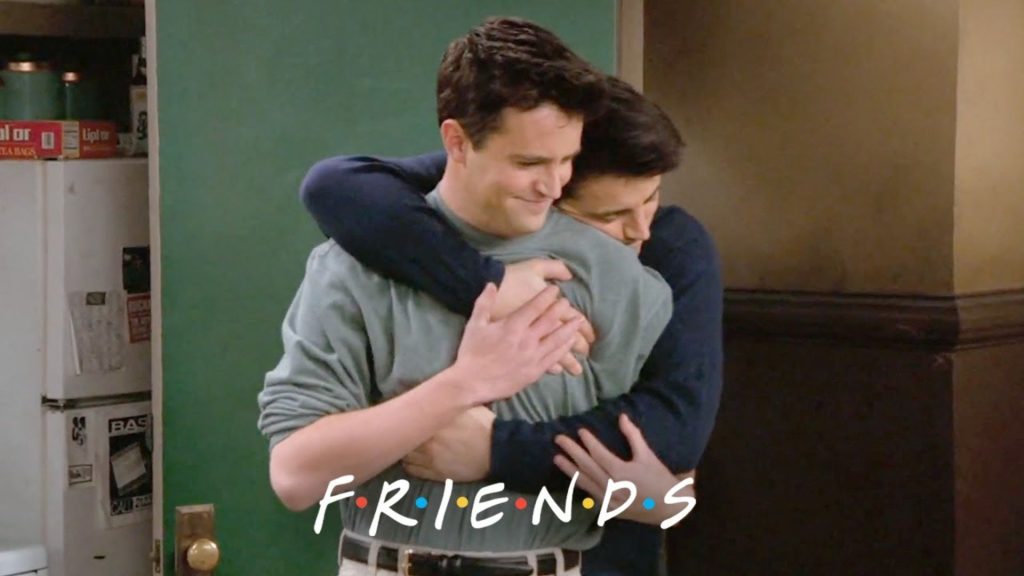 Joey hugging Chandler from behind at the entrance to their apartment. They smile as Chandler embraces Joey warmly. The logo of the show is centred beneath the two at the bottom of the image.