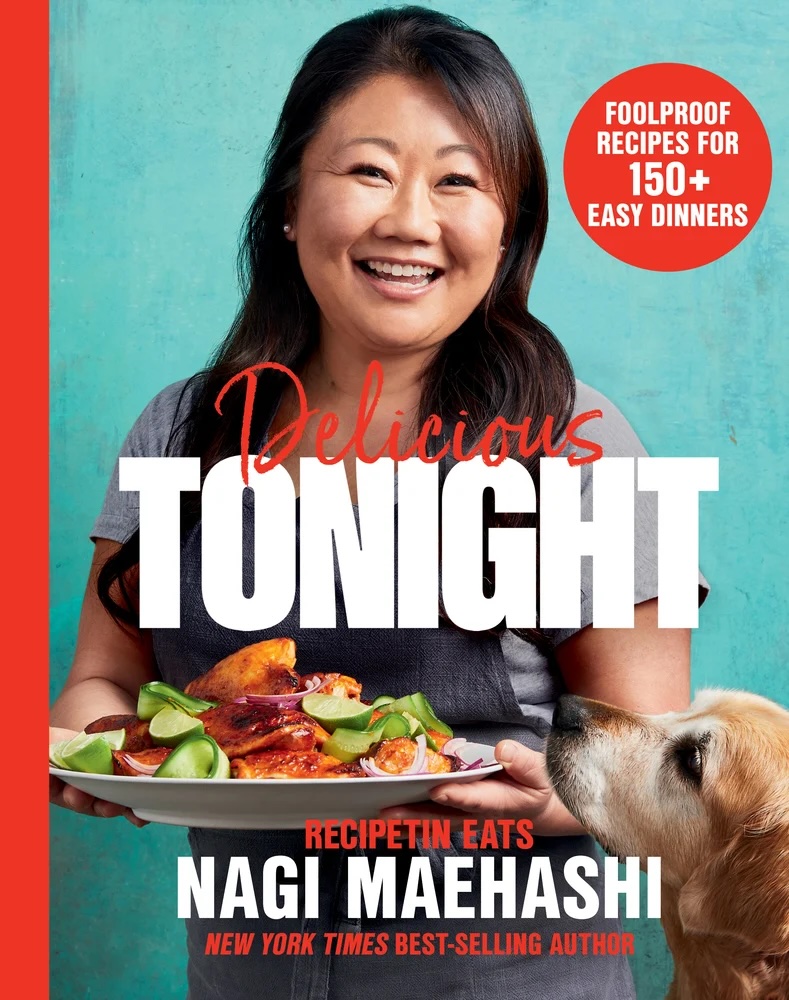 Delicious Tonight by Nagi Maehashi book cover, depicting Maehashi smiling at the camera in a grey shirt, carrying a plate of food with a dog sniffing it. The title is written in white, bold text while some of the smaller text is in red.
