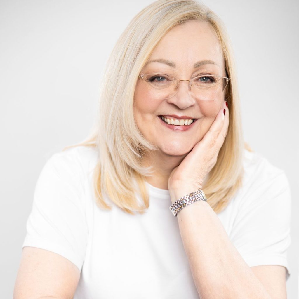 Author Ramona Baillie smiles in front of a white background as she rests her cheek on her hand. She has collarbone-length platinum blond hair and wears thin gold wire-framed glasses, a white T-shirt, and a silver watch.