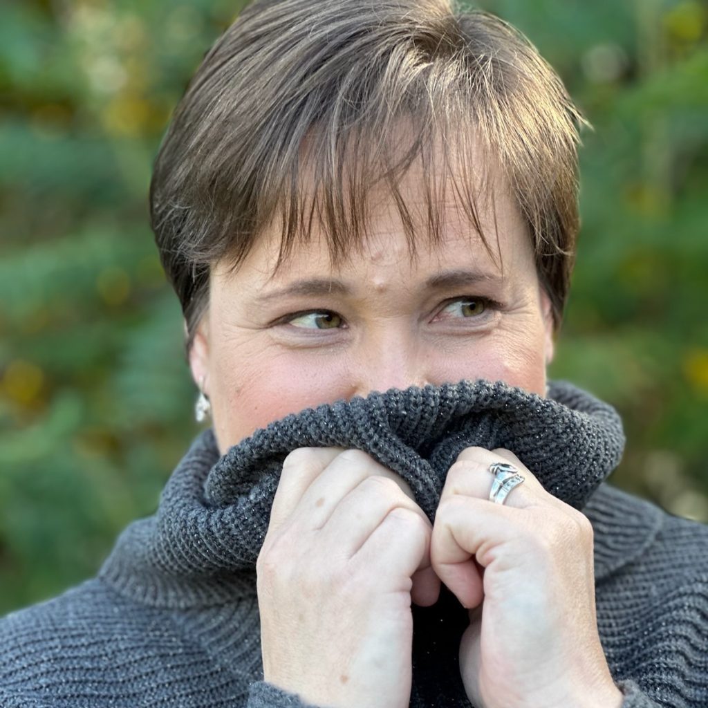 Brigid Barry, a light-skinned woman with short brown hair, pulls up her turtleneck over her mouth as she looks off to the viewer's right. Tree foliage can be seen out of focus behind her.