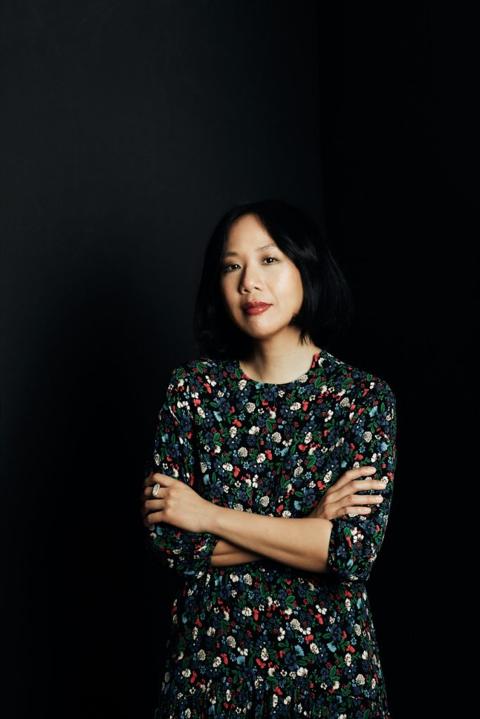 Jen Sookfong Lee, a Chinese Canadian woman with short black hair and a long-sleeved flower print dress, stands with her arms crossed in front of a black background, looking at the viewer.