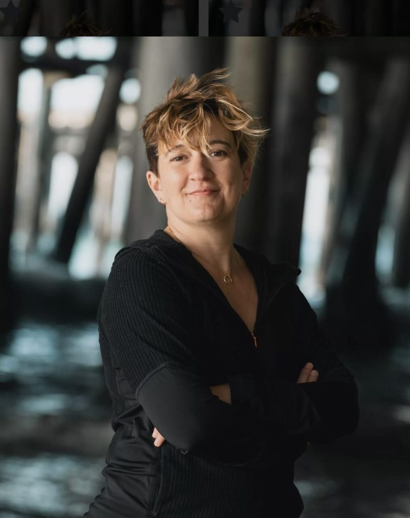Kathleen Kaufman, a light-skinned woman with short brown and blonde hair, stands with her arms crossed; the background is out of focus.