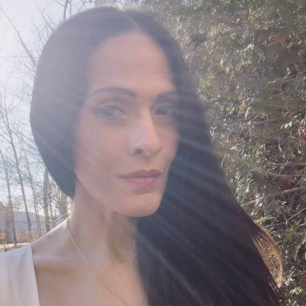 Cynthia Pelayo, a light-skinned woman with long dark hair, stands in front of a group of trees, rays of sun streaming from behind her.