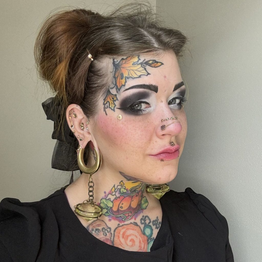 Rikki Goodwin, a light-skinned woman with freckles, piercings, and fall-themed tattoos on her temple and neck, stands in the corner of two off-white walls, facing the viewer at an angle to their right. Her hair is brown and tied up with a black bow, and she wears black and white eyeshadow and a black shirt.