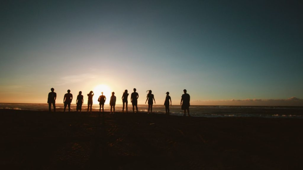 Eleven people standing on a dark beach during early sunrise. Their silhouettes are illuminated by the sun rising above the sea line into a cloudless light blue sky
