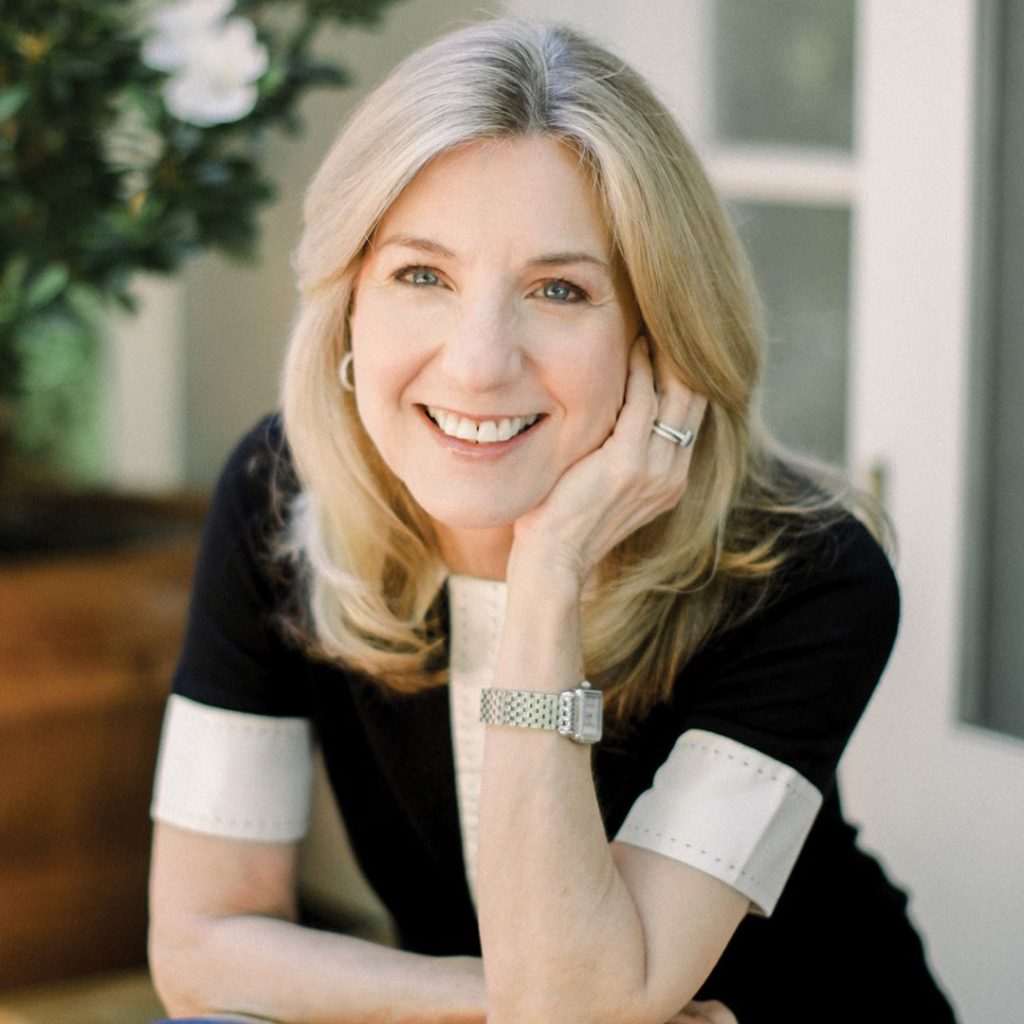 Author Carla Malden smiles and rests her left cheek on her palm. She has collarbone-length blonde hair, fair skin, and blue eyes and wears a black t-shirt with white trim around the sleeves, a silver watch with a rectangular face, a silver wedding band, and small silver hoop earrings.