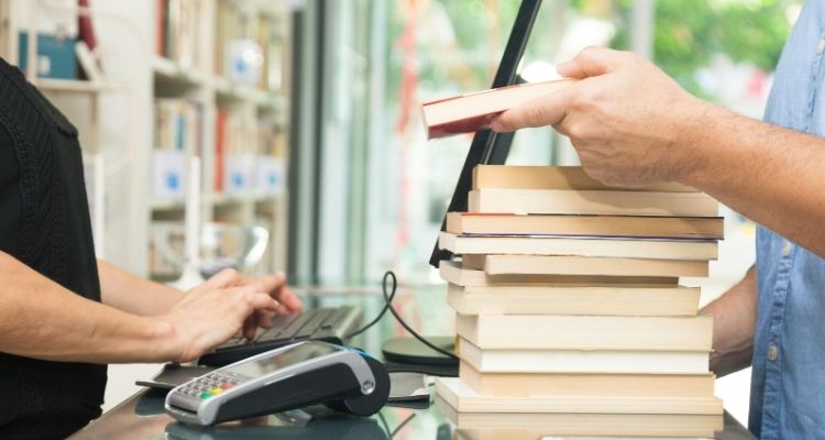 Depicts a person's hands buying a stack of books and a cashier's hands checking them out.