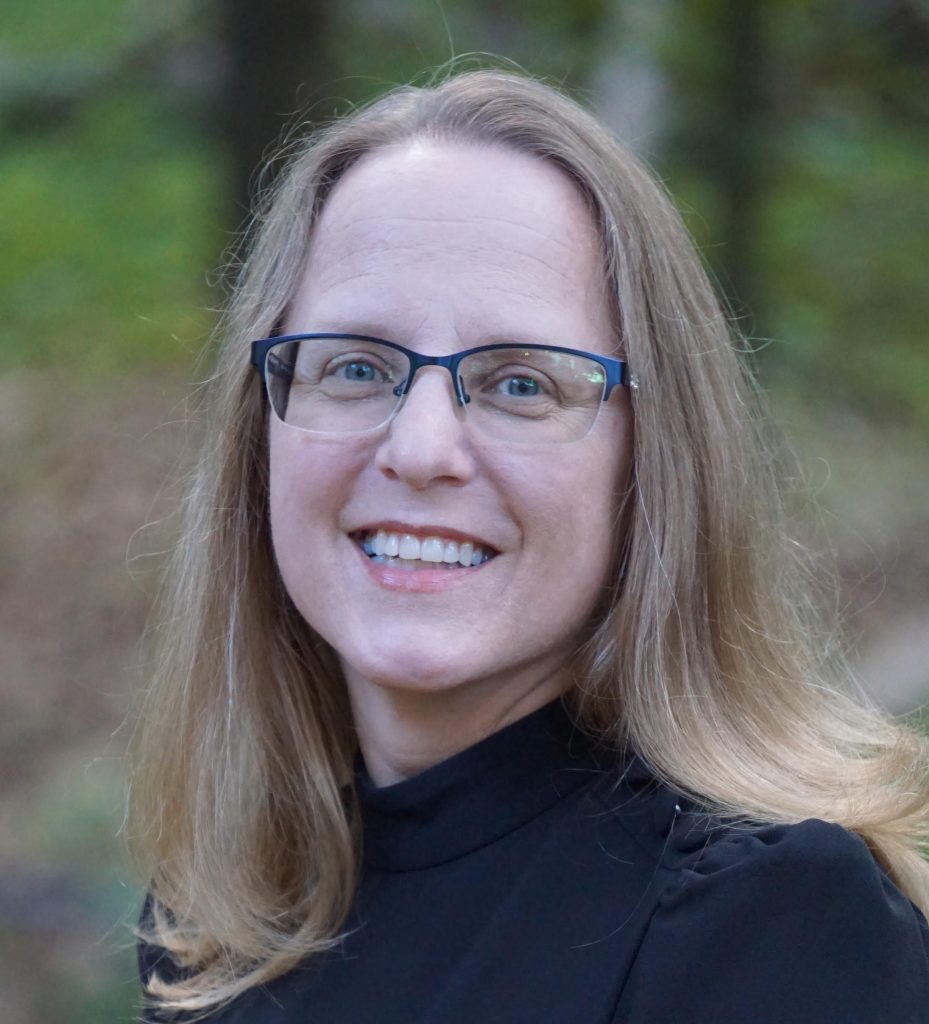 Stacy S. Jensen, a light-skinned woman with long blonde hair, glasses, and a black top, smiles at the viewer. Trees can be seen out of focus behind her.