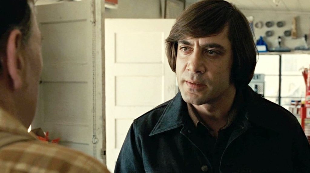 A still from No Country for Old Men showing Anton Chigurh, played by Javier Bardem, staring intently at a gas station clerk during a tense scene.
