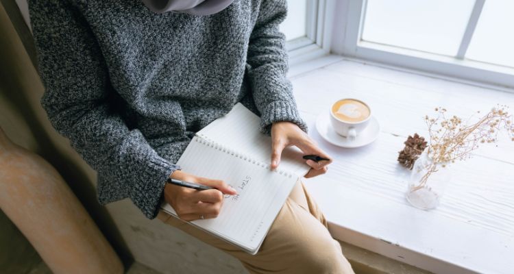 Depicts a woman sitting on a windowsill writing in a journal with a cappuccino and a plant beside her.