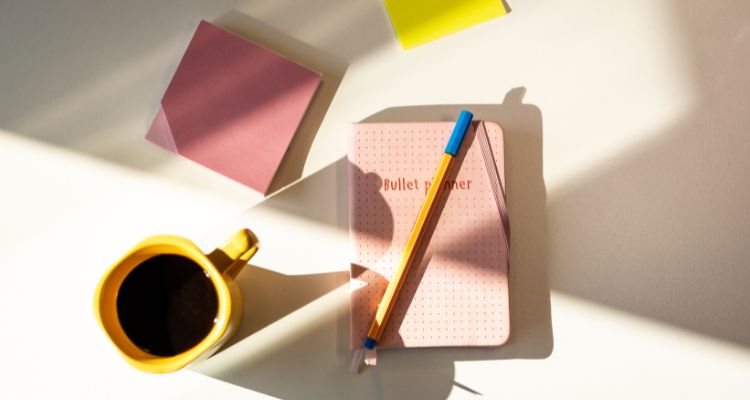 Depicts a pink bullet planner with a pencil, pink sticky notes, and a yellow mug of coffee beside the planner.