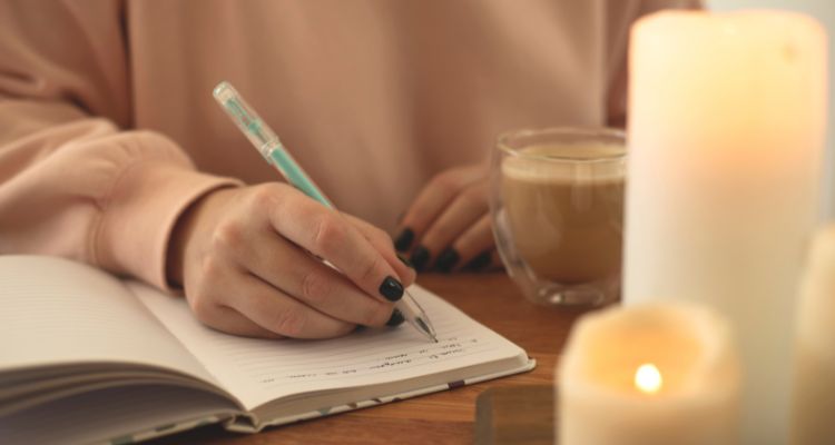 Depicts a woman's hands as she writes in a journal with a coffee and lit candles beside her.