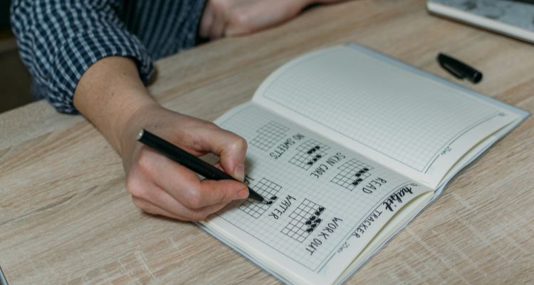 Depicts a hand filling out a habit tracker in a journal, tracking habits like reading, working out, water, and skin care.